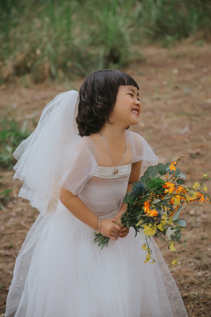 Hình ảnh cô dâu nhí cầm bó hoa màu vàng cam đang cười rất hồn nhiên và đáng yêu