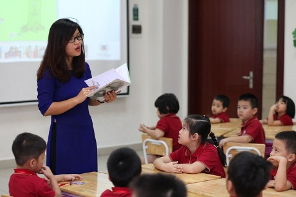 Cô giáo mặc áo dài màu xanh đang cầm sách giảng bài cho học sinh