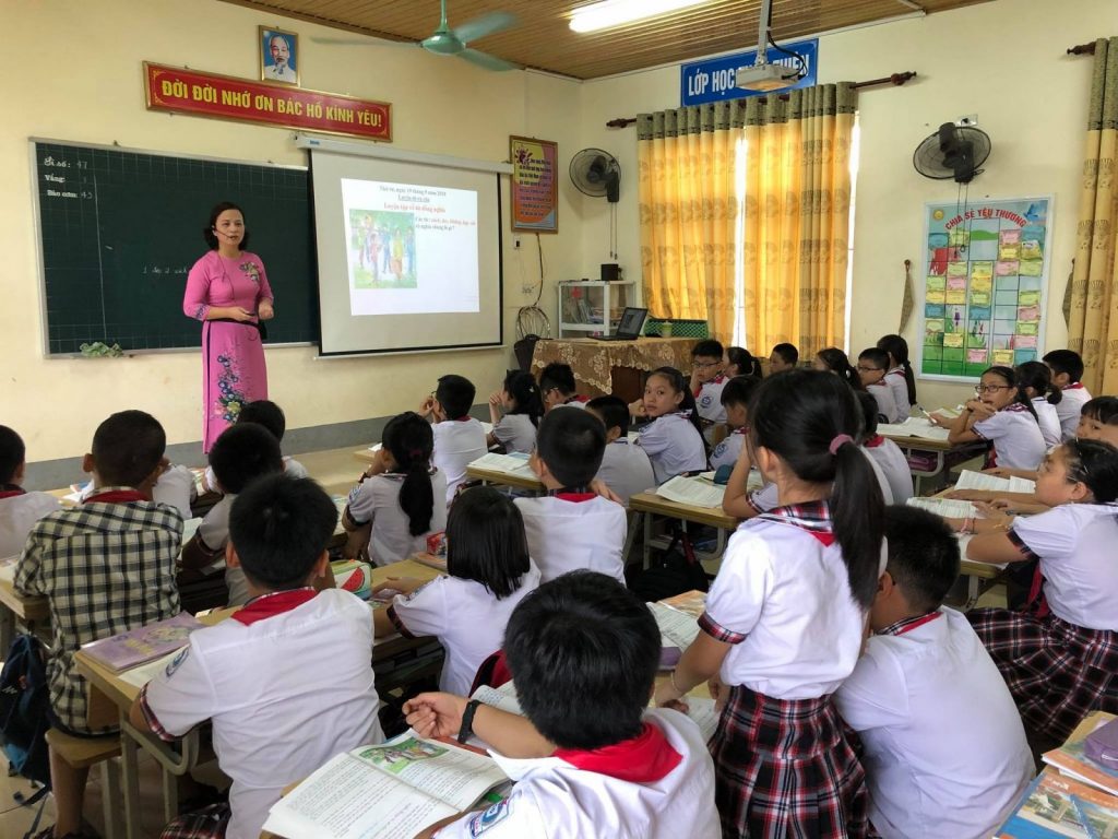 Cô giáo mặc áo dài màu hồng đang đứng trên bục giảng bài cho học sinh