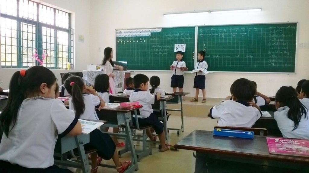 Cô giáo đang lắng nghe hai em học sinh lên bảng phát biểu bài